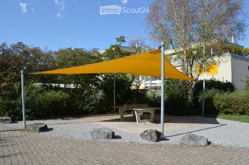 Outdoor seating area with a yellow canopy, picnic table, and scattered rocks on a paved surface.