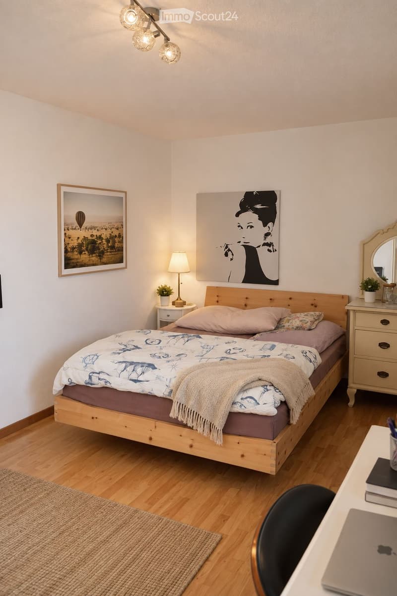 Cozy bedroom with a wooden bed, nightstand, lamp, and artwork on the walls. A small desk with a laptop and chair is visible, along with a mirror and plant.