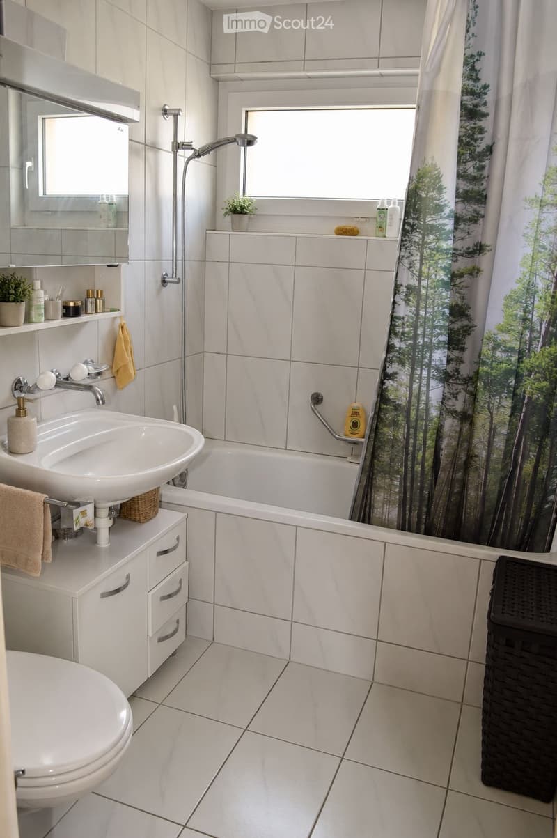 Modern bathroom with white tiles, a sink with a mirror cabinet, a toilet, a bathtub with a shower curtain, and a small plant on the windowsill.