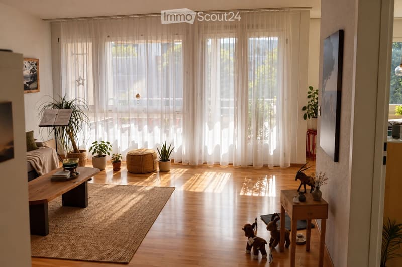 Bright living room with wooden flooring, large windows with sheer curtains, a sofa, wooden bench, ottoman, potted plants, and decorative items including toy animals.