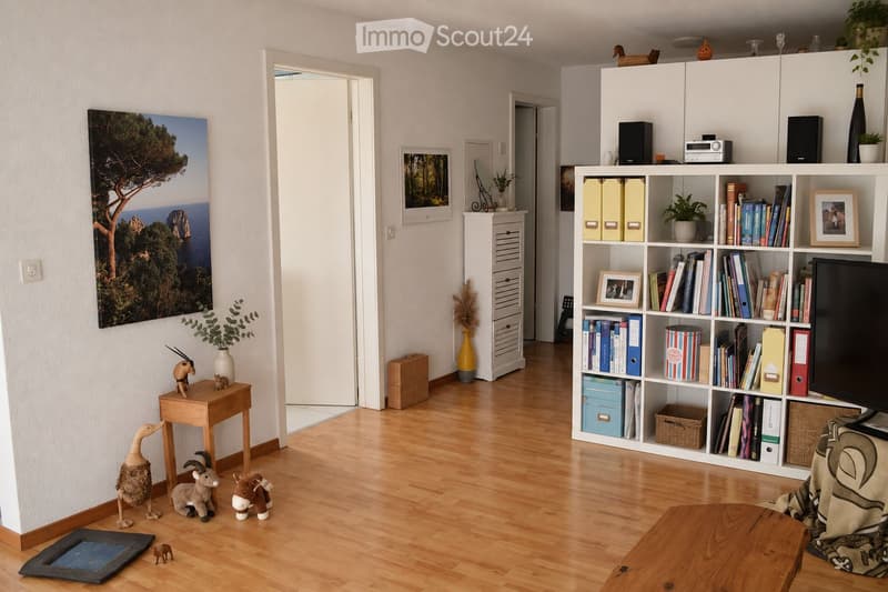 A well-lit living room with wooden flooring, a large bookshelf filled with books and decorative items, a small wooden table with toys, and a large painting on the wall.