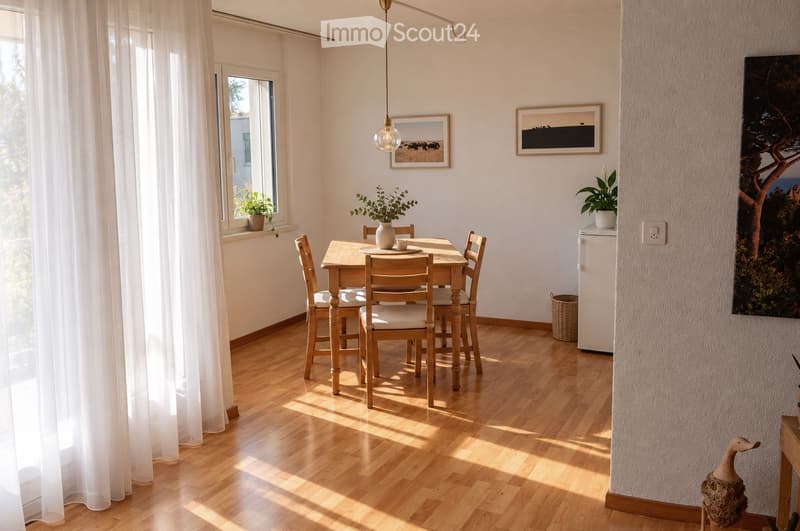 Bright dining area with wooden floor, large window with white curtains, dining table with chairs, vase with plants, picture frames on walls, small refrigerator, and decorative items.