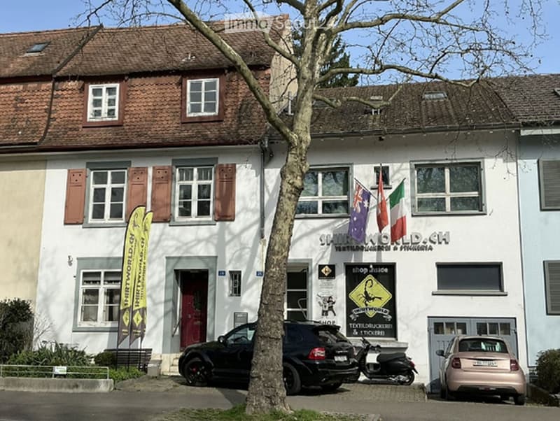 Two-story building with white facade, multiple windows, and a tiled roof. Flags of Australia, Italy, and Switzerland are displayed, and there is signage for "Shirtworld.ch".