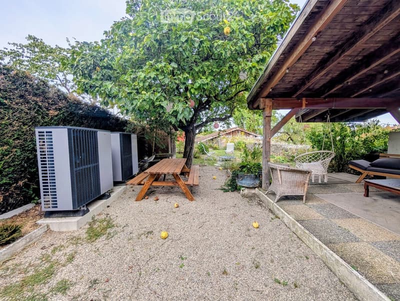 Outdoor patio area with a wooden picnic table, wicker chair, sun lounger, and air conditioning unit, surrounded by greenery and a tree.