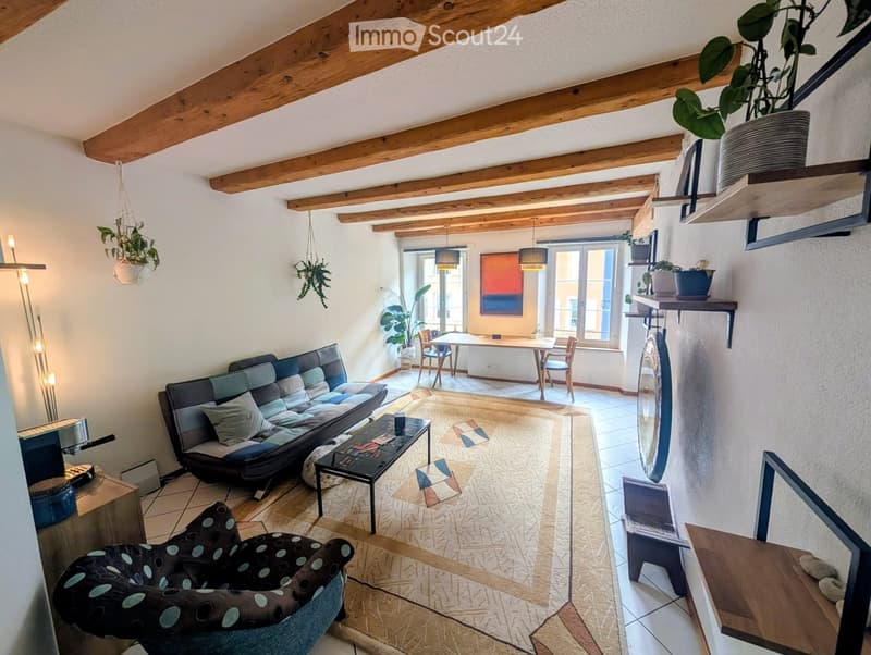 Living room with exposed wooden beams, a sofa, coffee table, armchair, dining table, and shelves with plants.