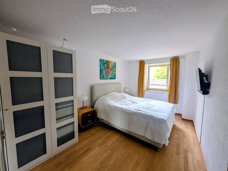 A well-lit bedroom with a wooden floor, a large bed with white bedding, a nightstand with a lamp, a painting on the wall, a wardrobe with mirrored doors, and a television on a stand.