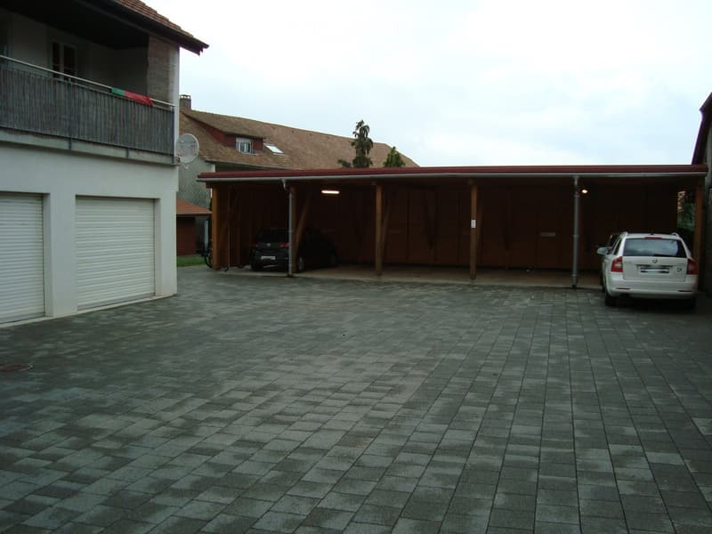Garage with two cars parked, one under the roof, another in the open, white garage doors, brick building, paved parking lot