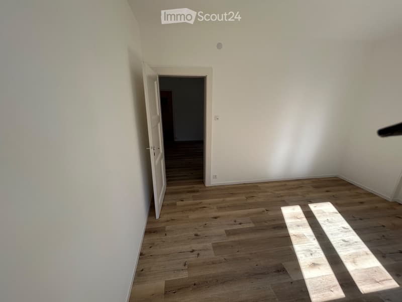 Empty room with light wooden flooring and white walls, featuring an open doorway leading to another room.