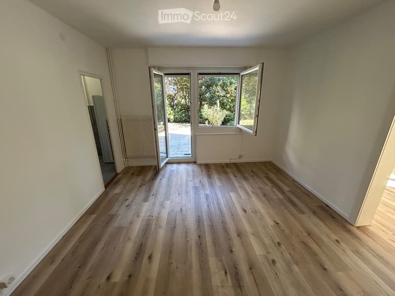 Empty room with light wooden flooring, white walls, and large windows with open shutters leading to an outdoor area.