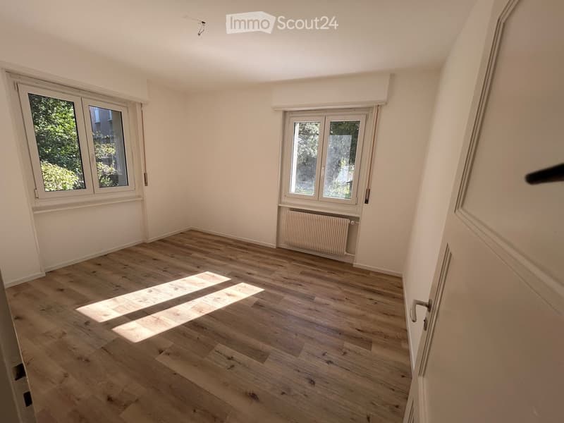 Empty room with wooden flooring, white walls, and large windows allowing natural light to enter. There is a radiator under one window and a door visible on the right side.