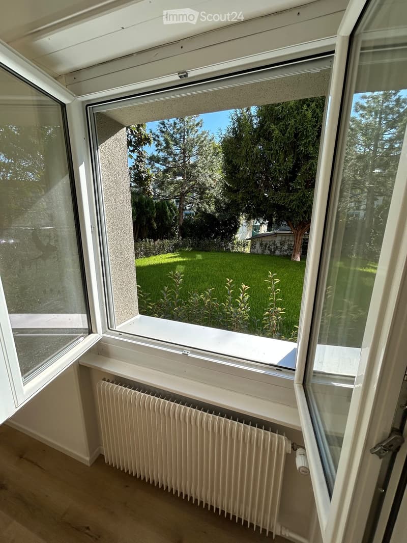 Empty room with wooden flooring, white radiator, and large window offering a view of a green garden and trees.