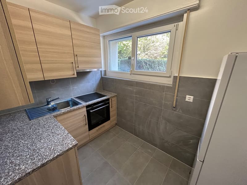 Modern kitchen with light wooden cabinets, granite countertop, stainless steel sink, and built-in oven. Grey tiled backsplash and floor.