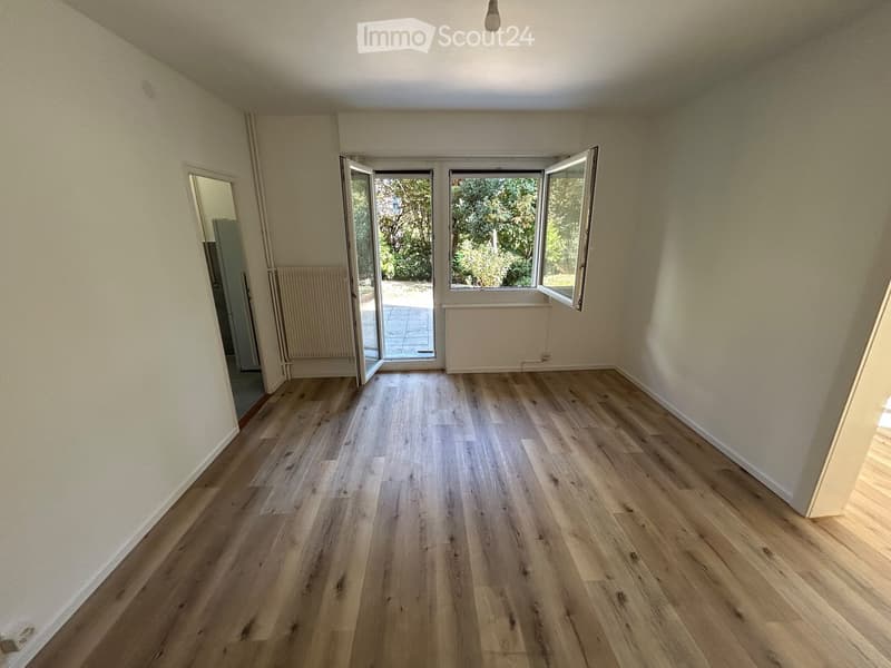 Empty room with light wooden flooring, white walls, and large windows with open shutters leading to an outdoor area.