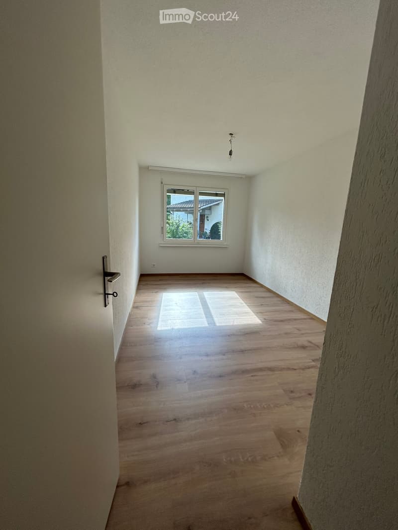 Empty room with wooden flooring, white walls, and a window with a view of greenery outside.