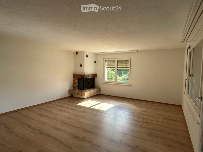 Empty room with wooden flooring, white walls, and a fireplace with a brick surround. A window with blinds allows natural light to enter.