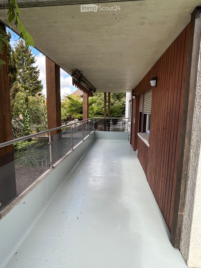 Modern balcony with wooden walls, glass railing, and a smooth concrete floor, featuring outdoor lighting and a view of greenery.