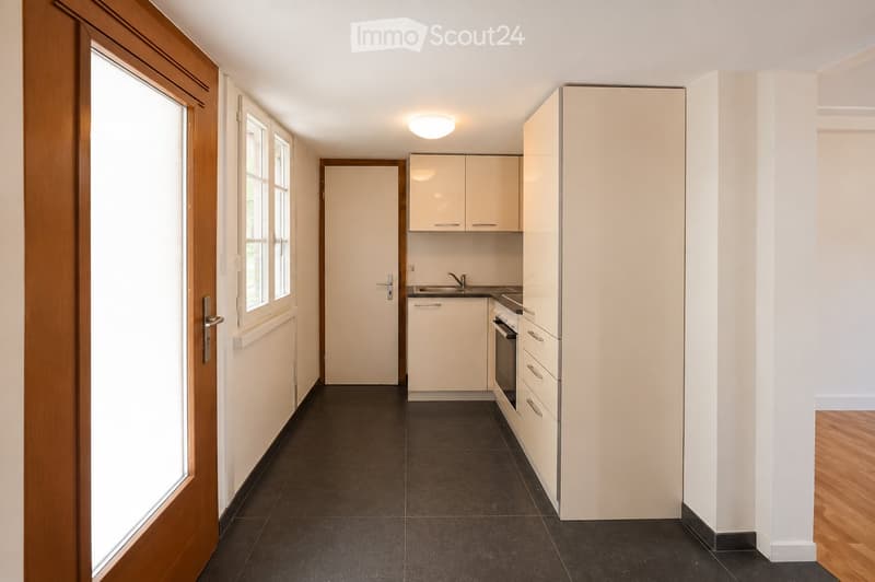 Modern kitchen with white cabinets, black tiled floor, sink, faucet, and oven. The space is well-lit with a ceiling light and has a wooden door leading to another room.