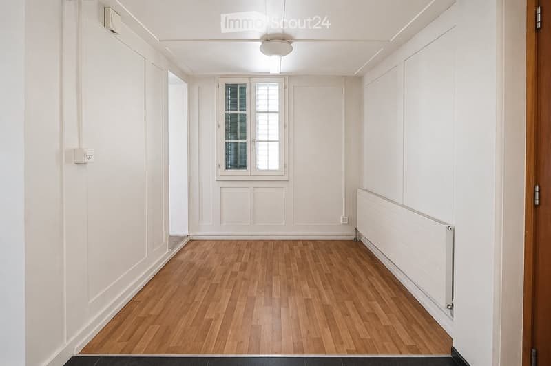 Empty hallway with wooden flooring, white walls, and a window with blinds. No furniture or equipment present.