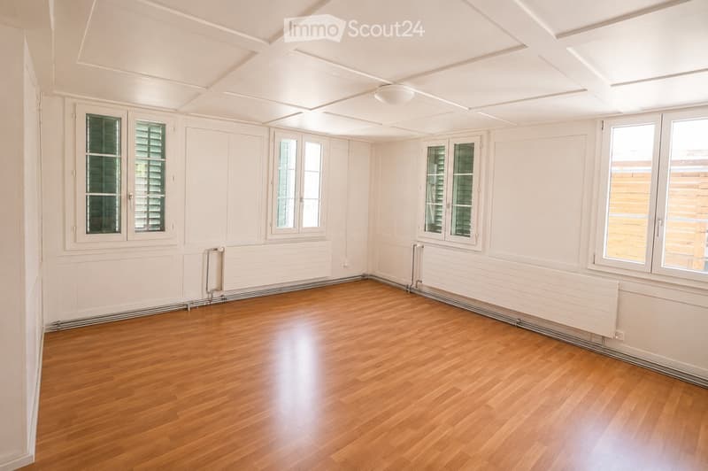 Empty room with wooden flooring, white walls, multiple windows with shutters, and radiators along the walls.