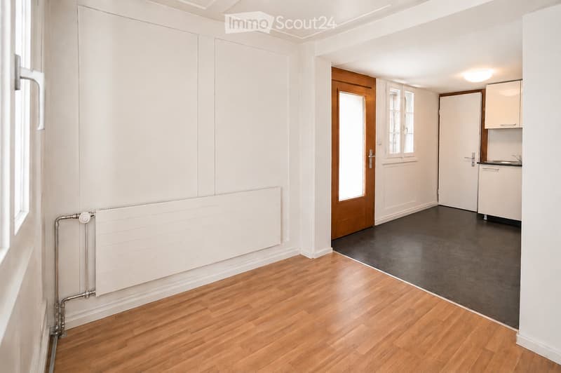 Empty room with wooden flooring, white walls, radiator, and a doorway leading to another room with a small kitchen area.