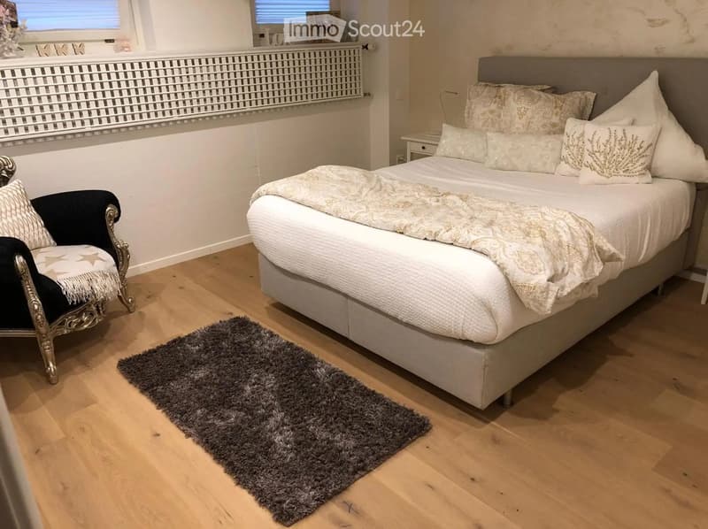 Cozy bedroom with a large bed, patterned pillows, a decorative throw, a plush grey rug, and a stylish black and gold armchair.