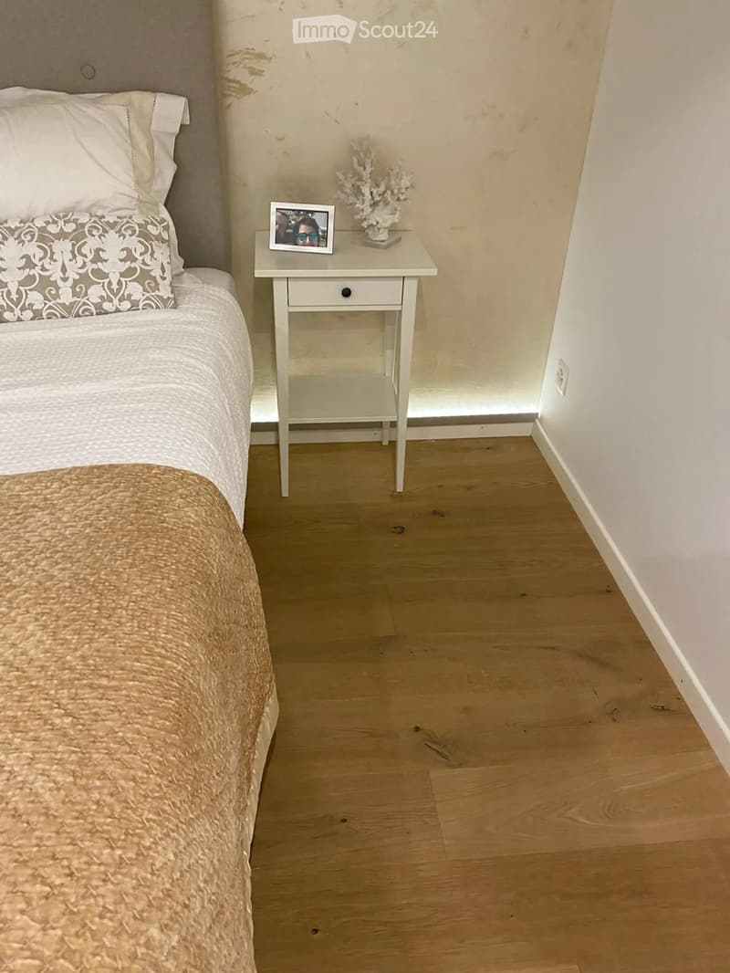 Cozy bedroom with a neatly made bed, a nightstand with a picture frame and decorative item, and light wooden flooring.