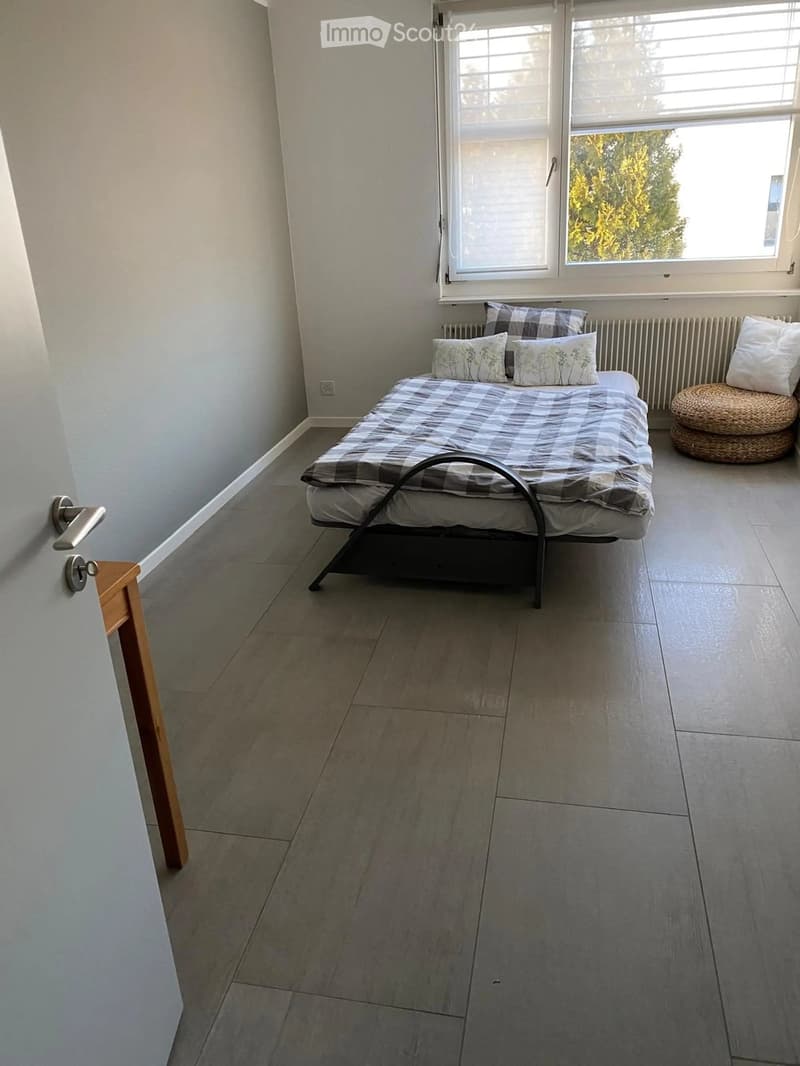 A small bedroom with a single bed, pillows, a wicker stool, and a window with blinds. The room has light-colored walls and a tiled floor.