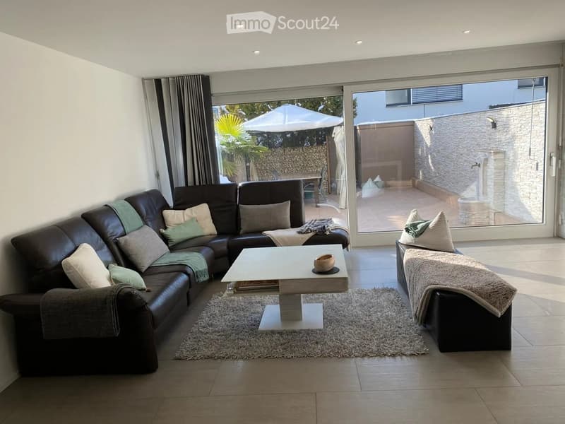 Modern living room with a dark leather sectional sofa, a white coffee table, and a shaggy rug. Large glass doors open to a balcony with outdoor furniture.