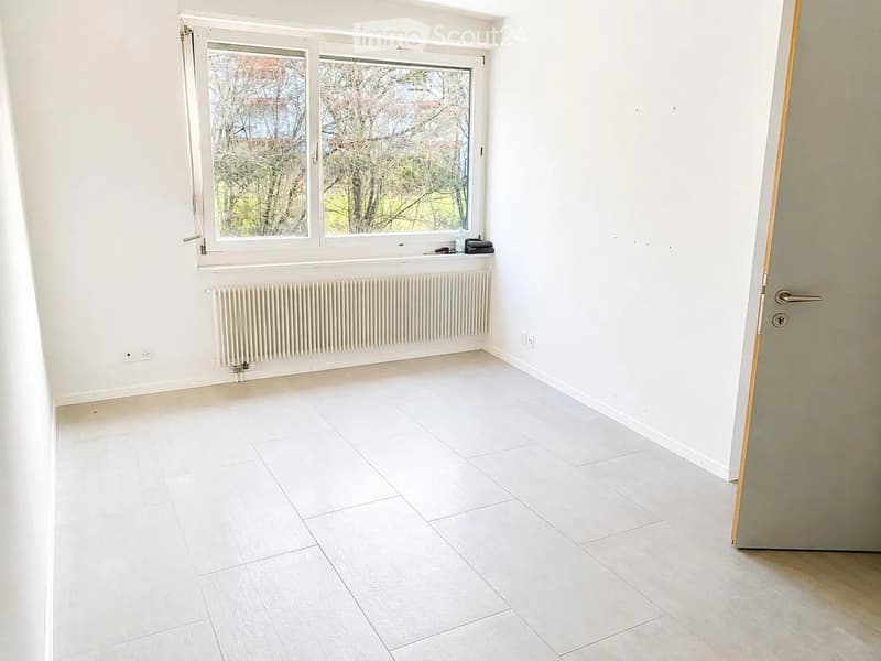 Empty room with white tiled floor, white walls, large window with view of trees, radiator, and a door.