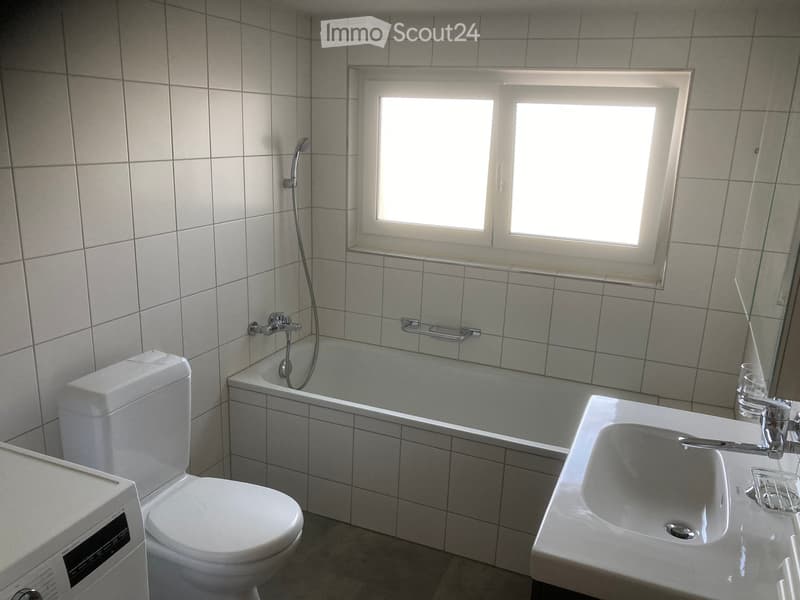 Modern bathroom with white tiles, a bathtub, toilet, sink, and a window above the bathtub. Includes a shower hose and a mirror on the wall.