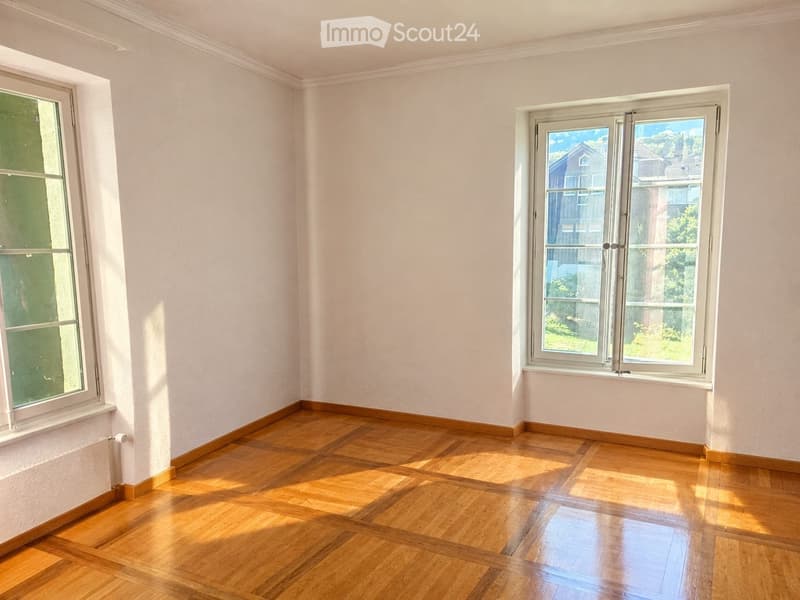 Empty room with light-colored walls and wooden flooring, featuring two windows with white frames and a radiator visible on the left side.