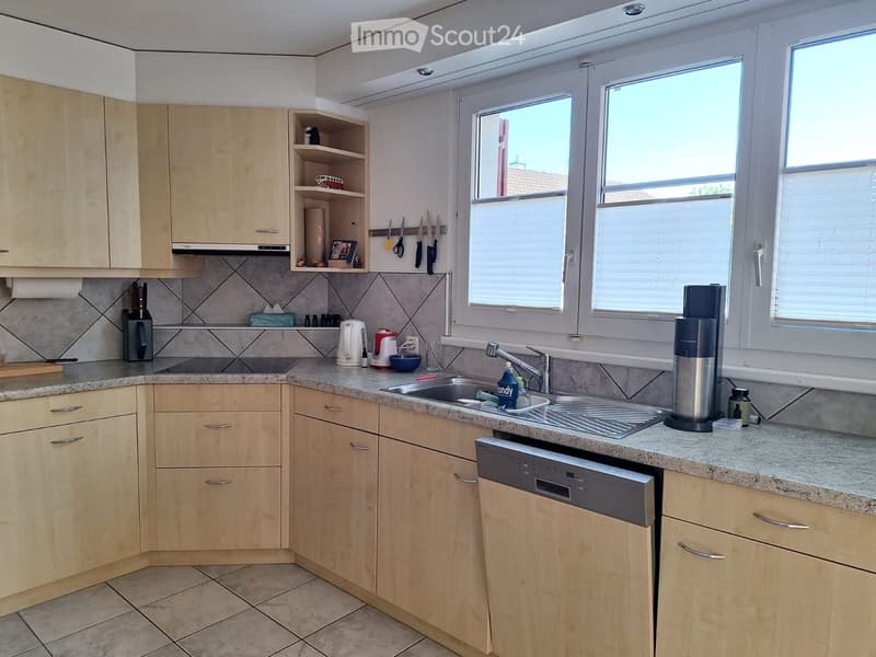 Modern kitchen with light wooden cabinets, granite countertop, stainless steel sink, and various appliances including a blender and toaster. The kitchen features a window with blinds and a tiled backsplash.