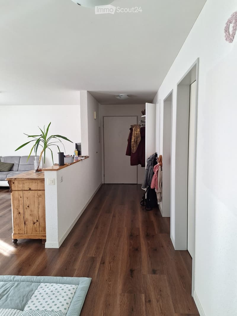 Bright hallway with wooden flooring, white walls, and a coat rack with jackets. A small wooden console table holds a plant and other items, and a pet bed is visible on the floor.