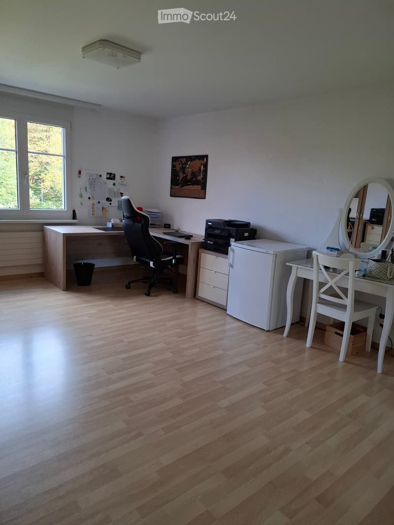 Well-lit room with wooden flooring, featuring a large desk with a chair, a printer, a small refrigerator, a vanity with a mirror, and a small table with a chair.