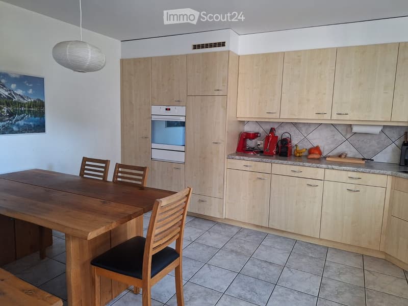 Bright kitchen with light wooden cabinets, granite countertops, a wooden dining table with four chairs, and various appliances including a coffee maker and a fridge. A picture of a mountain landscape is on the wall.
