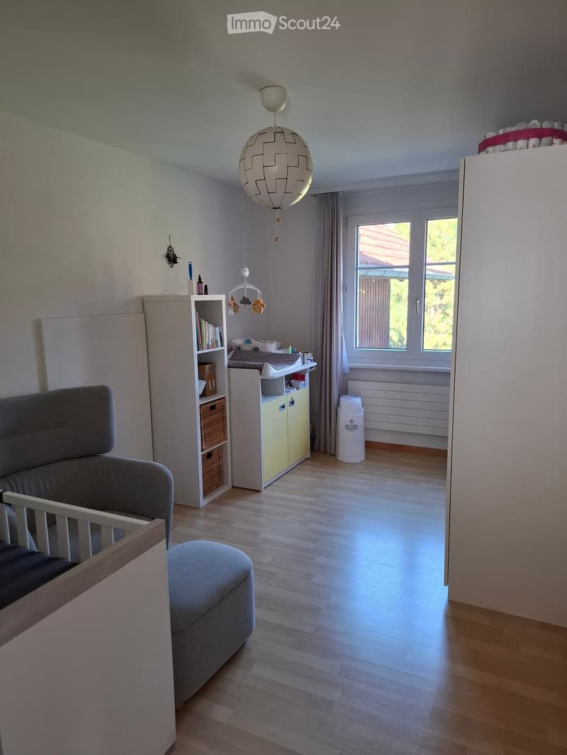 Brightly lit room with wooden flooring, a grey sofa, ottoman, bookshelf, baby chair, and a small kitchen area with a bottle warmer.