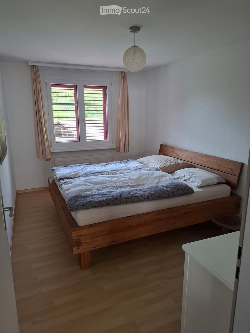 A well-lit bedroom with a wooden bed, pillows, a headboard, and a hanging light fixture. The room features wooden flooring and a window with curtains.