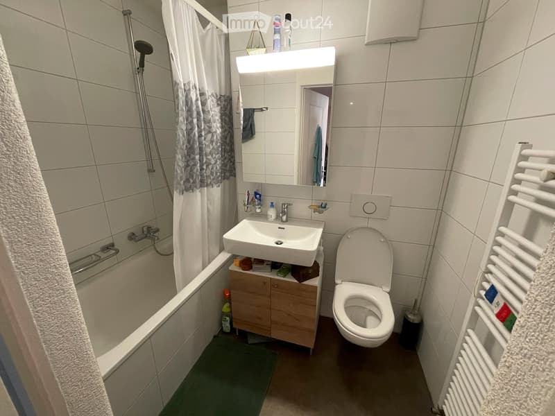 Modern bathroom with a white sink, toilet, bathtub, and a large mirror with built-in lighting. Includes a wooden cabinet under the sink, a white radiator, and a towel on the side.