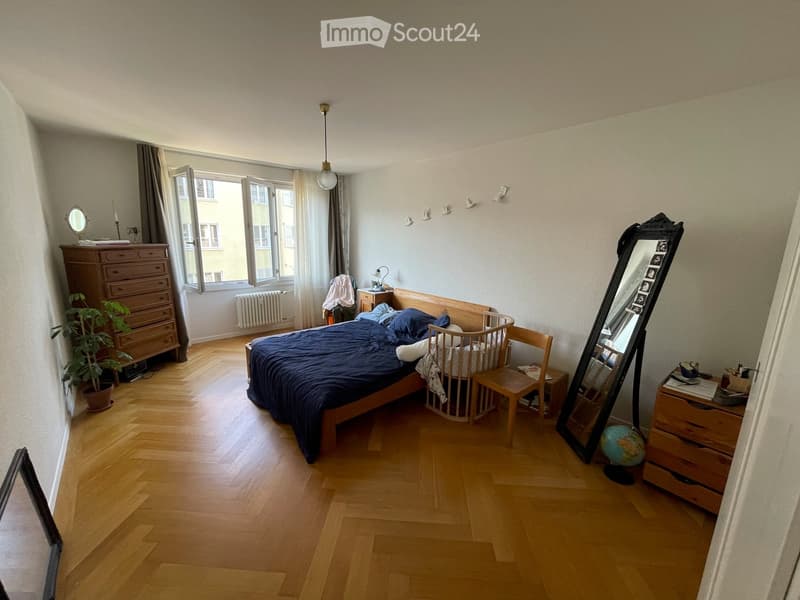 Cozy bedroom with wooden flooring, large window with curtains, bed with dark blue bedding, nightstand, mirror, chair, small table, dresser, plant, and a baby cradle.