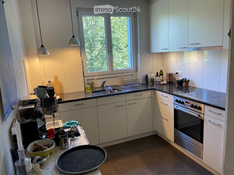 Modern kitchen with white cabinets, stainless steel appliances, and a large window. The counter is cluttered with various kitchen items including a coffee maker, bowls, a frying pan, and a dish rack.