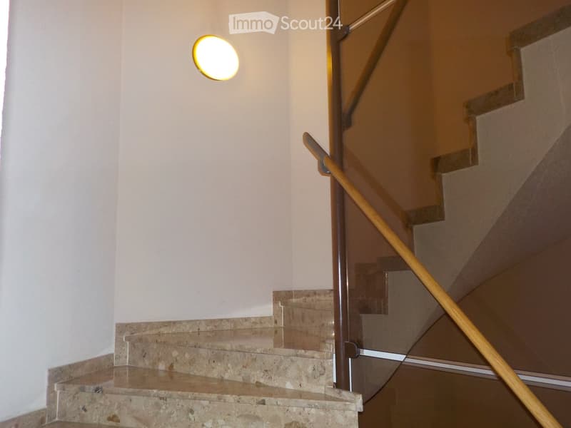 Staircase with beige marble steps, glass railing, wooden handrail, and a round wall-mounted light fixture.