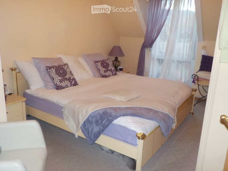 Cozy bedroom with a large bed, decorative pillows, a nightstand with a lamp, and a small chair near the window.