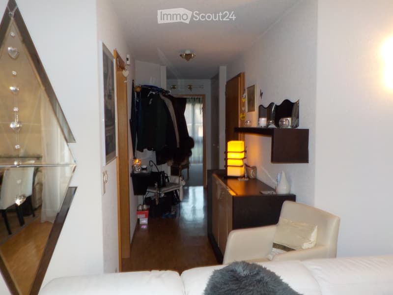 A well-lit hallway leading to a furnished room with a white sofa, a chair, a small table, a mirror, and a coat rack with jackets hanging on it.