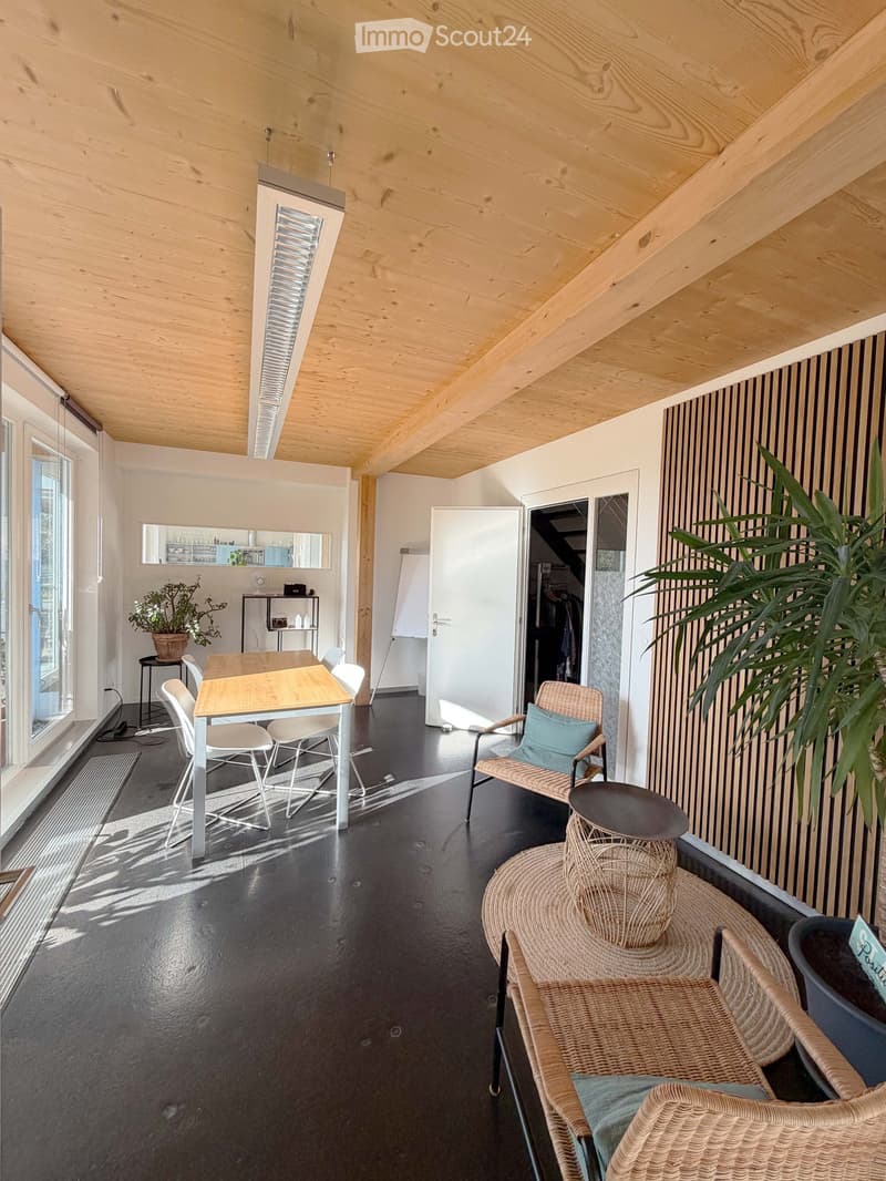 Modern, open-plan room with wooden ceiling beams, black flooring, and large windows. Features a dining table with chairs, a wicker seating area with a stool, and a potted plant near the window.
