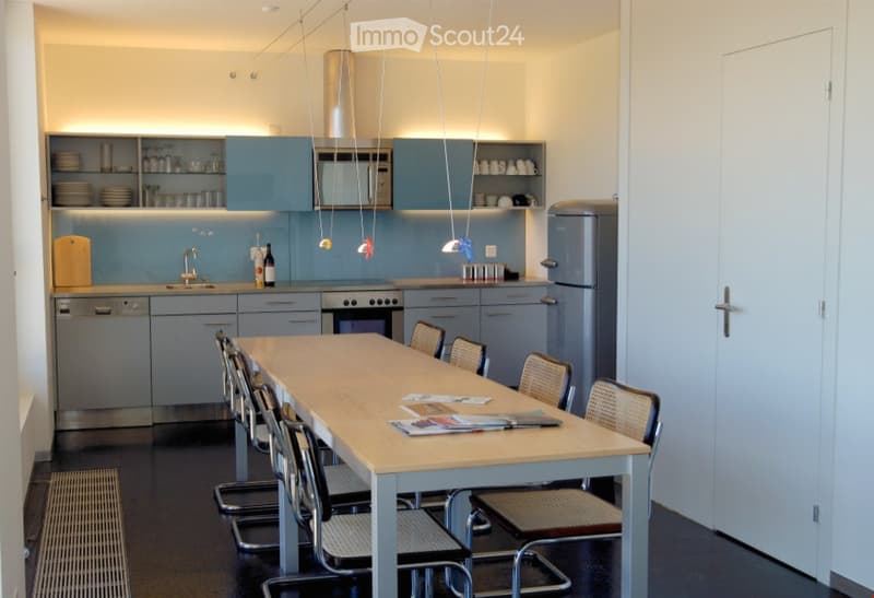 Modern kitchen with light blue cabinets, stainless steel appliances, a long wooden dining table surrounded by chairs, and various kitchen items on the counter and shelves.