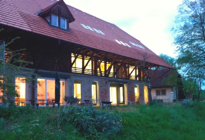 A large, two-story building with a red-tiled roof and wooden balcony, surrounded by greenery and trees.