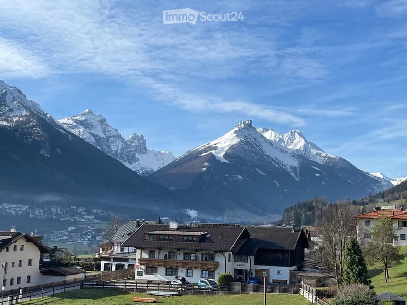 A large, multi-story house with a dark roof and white walls, situated in a picturesque valley surrounded by snow-capped mountains and a small town in the background.
