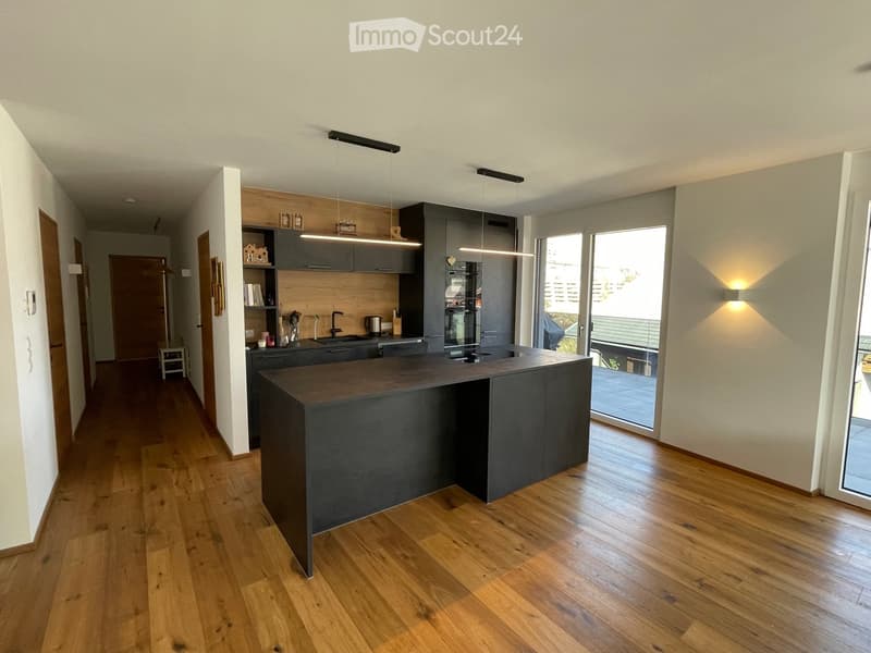 Modern kitchen with dark grey cabinets, a central island, wooden flooring, and built-in appliances. Features pendant lighting and a large window with a view of a balcony.
