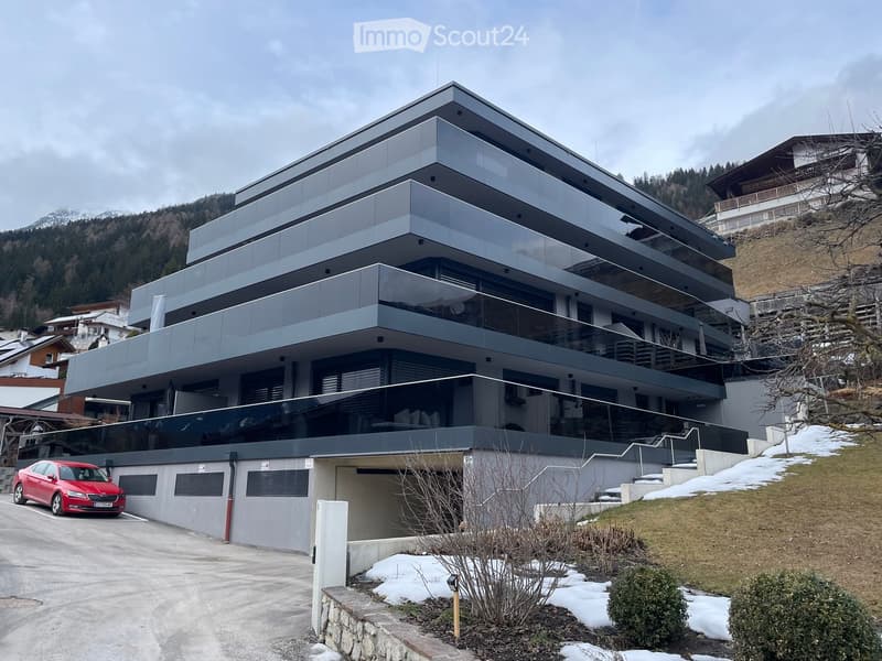 Modern multi-story house with large glass windows and balconies, situated in a scenic mountainous area with snow-covered ground and surrounding trees.