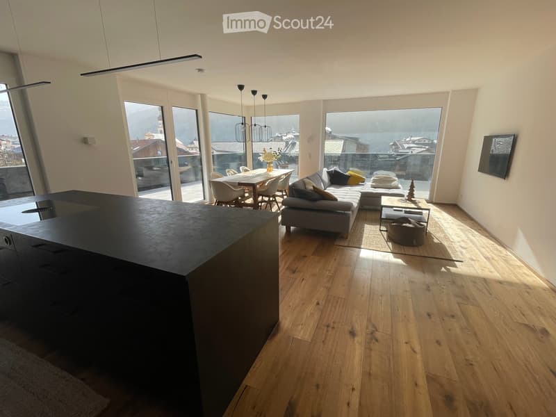 Modern living room with wooden flooring, large windows offering a scenic view, a grey sectional sofa, a coffee table, a dining area with a table and chairs, and a television mounted on the wall.